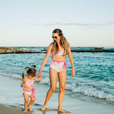 Mom and Daughter in Matching Tie dye swimsuits.  The toddler daughter is wearing a bikini with a bow.  The mom is holding her hand and wearing a matching high rise bikini in the same tie dye print. 